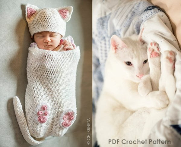 A baby in a cat themed crochet baby cocoon, hat, and booties; next to a curled up white cat.