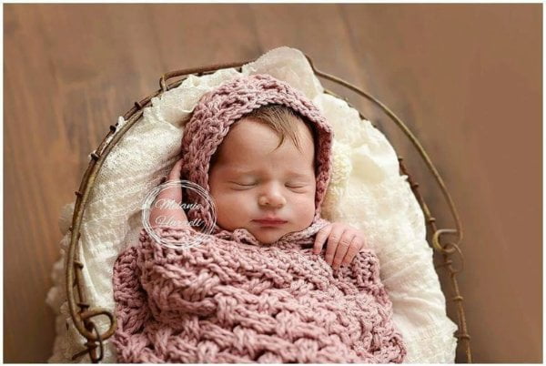 Newborn baby in pink bonnet, sleeping in a basket in a pink crocheted baby cocoon.