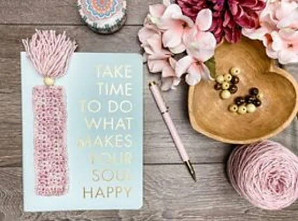 A pink crochet bookmark with tassels on a desk with flowers and a pen.