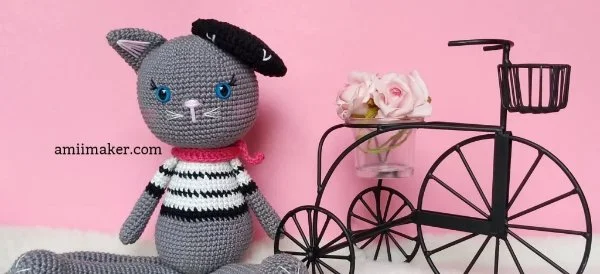 Crochet cat in a striped shirt wearing a beret and sitting next to a bicycle.