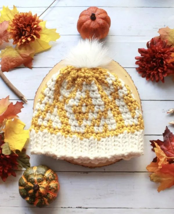 A yellow and white tapestry crochet beanie on a wooden board with autumn leaves.