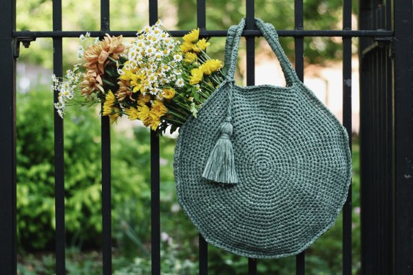 A green crochet circle bag with flowers hanging on a fence.