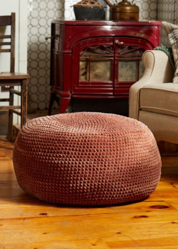 A rust-coloured crochet pillow pouf on a timber floor in a warm-looking living room.
