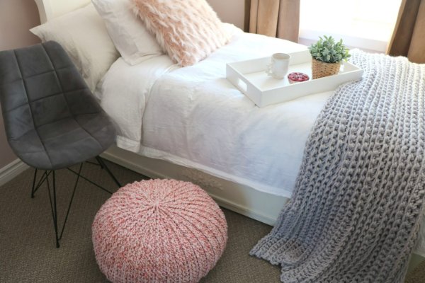 A pink crochet floor pouf next to a bed with a breakfast tray on it.