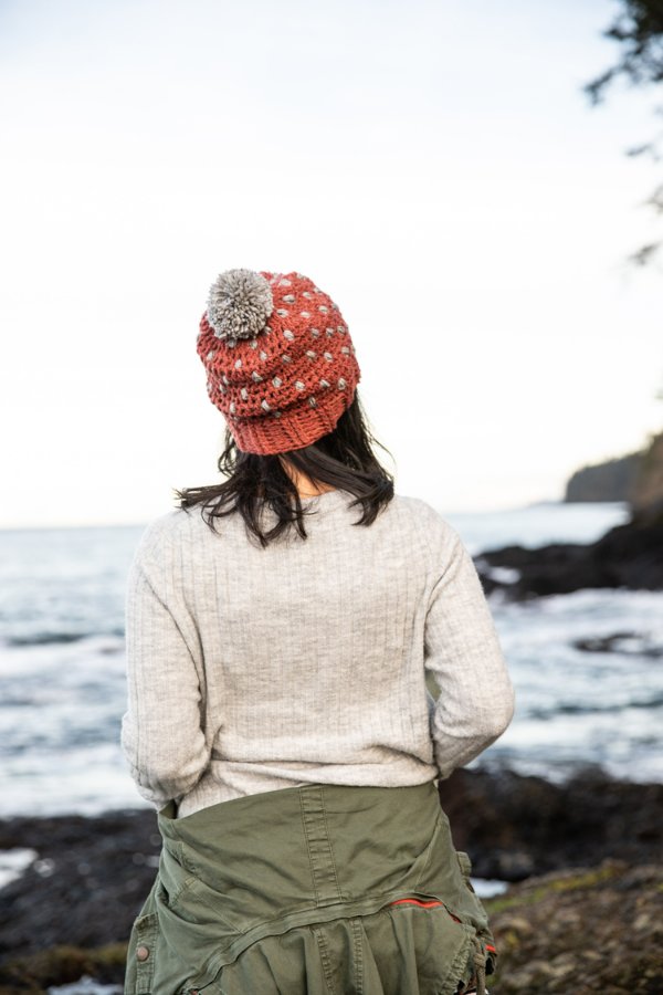 Back view of woman wearing a crochet beanie standing on rocky shore overlooking ocean.