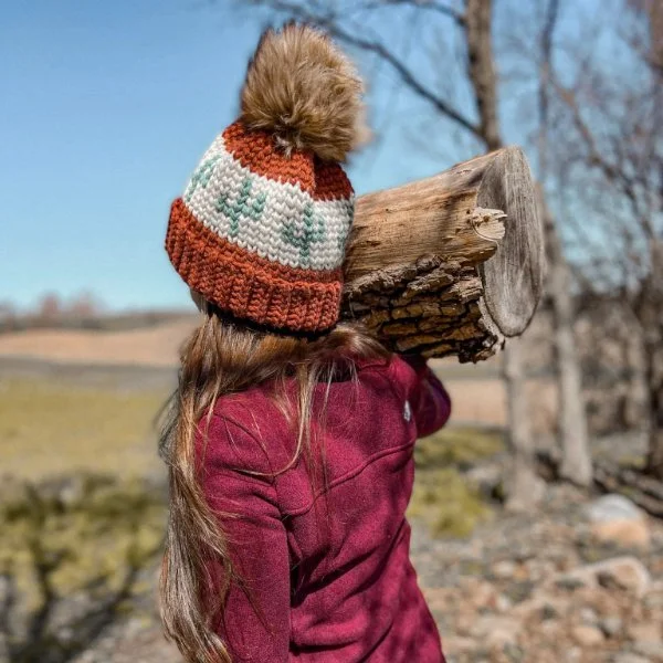 A woman wearing a tapestry crochet beanie carrying wood.