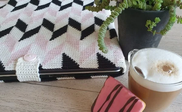 Black ,white, and oink tapestry crochet laptop sleeve with potplant and coffee.