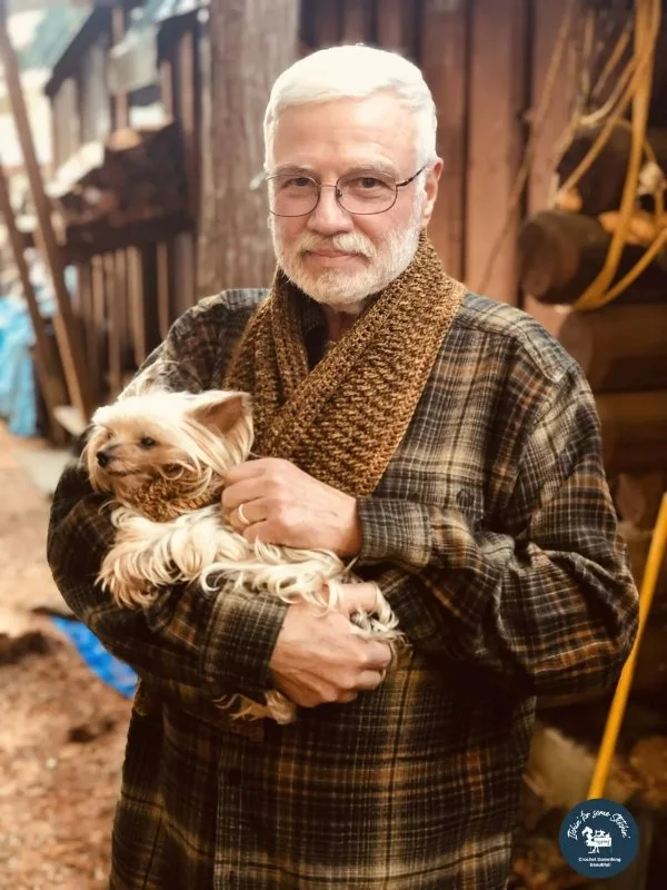 A man wearing a crochet scarf while holding a small dog.
