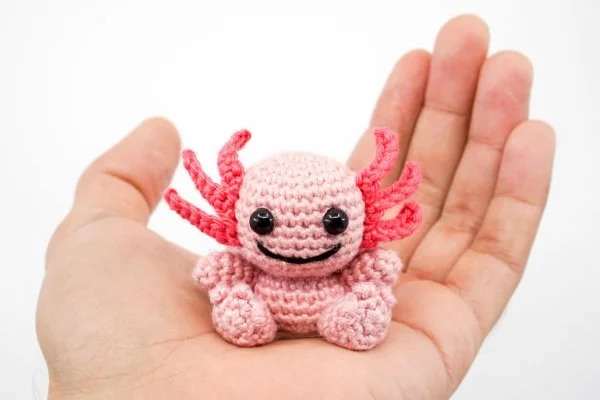 A small crochet axolotl sitting in the palm of a hand.