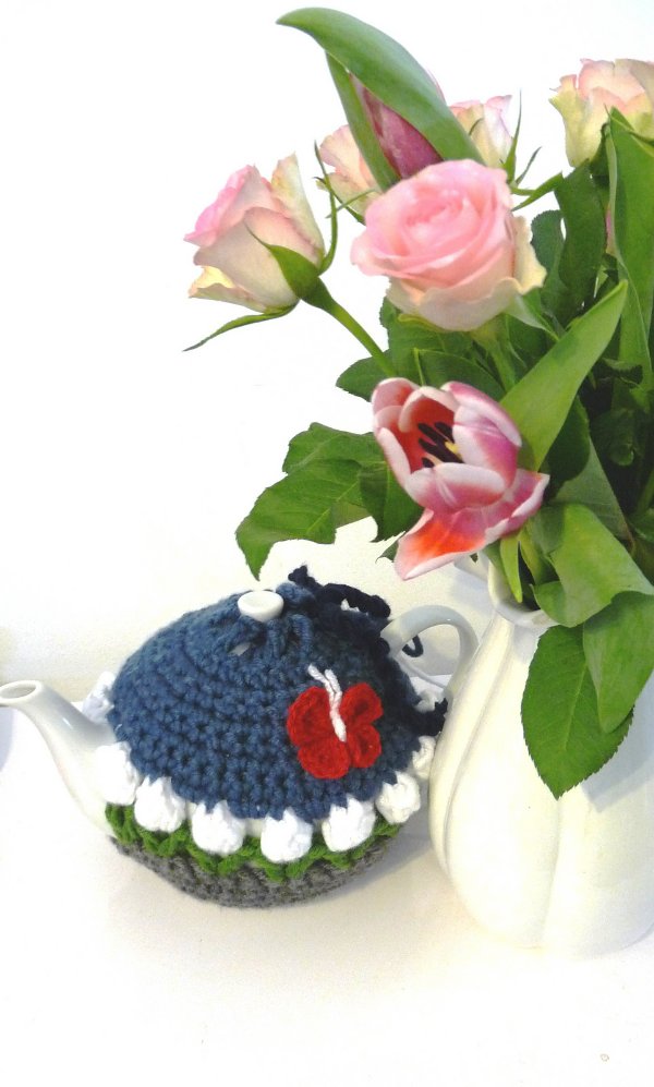 A teapot covered with a crochet tea cozy on a table with a bunch of flowers.