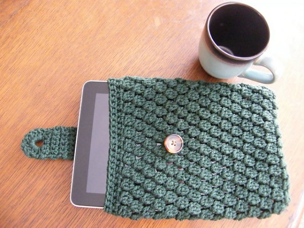 A green crochet tablet sleeve on a table with a coffee cup.