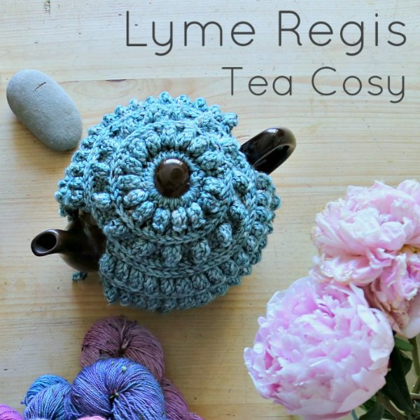 A black teapot with a blue crochet tea cozy, flowers and wool skeins.