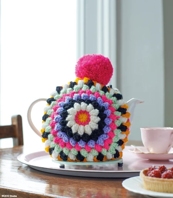 Brightly coloured crochet tea cozy with a pompom on a set tea table.