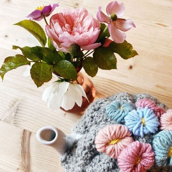 Crochet tea cozy with flower decoration on a table with a bunch of flowers.