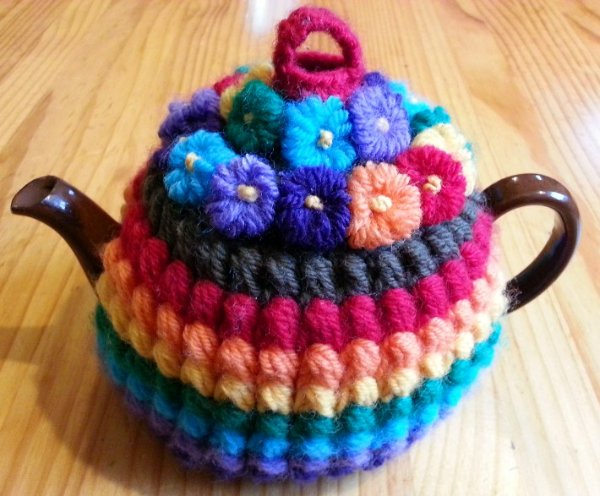 Rainbow-coloured crochet tea cozy with flower detail.