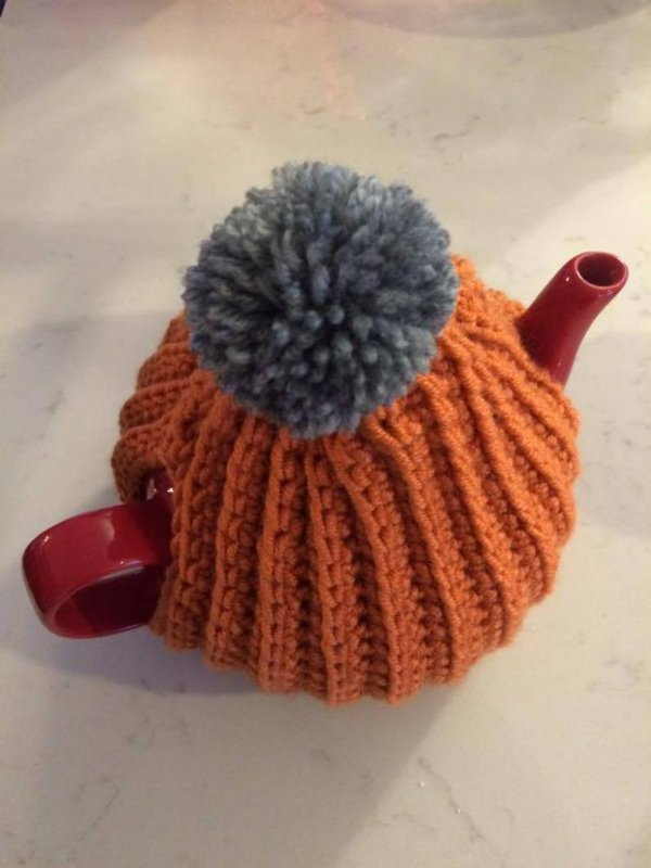 A read teapot in an orange crochet tea cozy with a grey pompom.