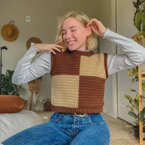 A woman sitting down in jeans and a checkerboard crochet sweater vest.