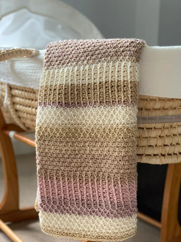A Tunisian crochet baby blanket in muted tones draped over a Moses basket.