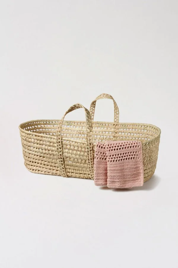 A filet crochet baby blanket hanging over a Moses basket.