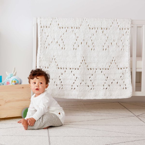 A baby sitting on the floor with a crib and white filet crochet blanket in the background.