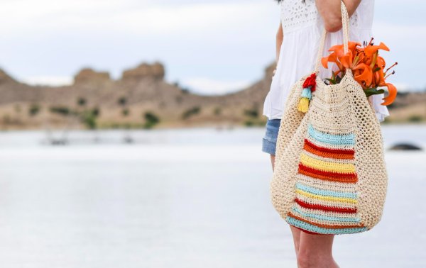 A woman holding a colourful crochet beach bag and orange lilies.