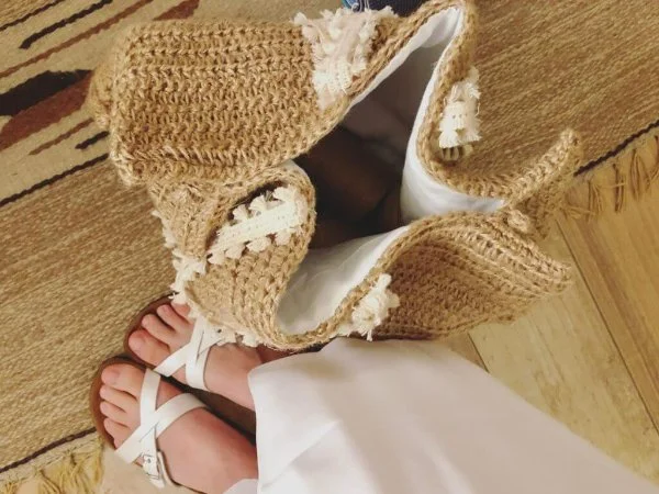 A womans feet and sandals next to a large jute crochet beach bag.
