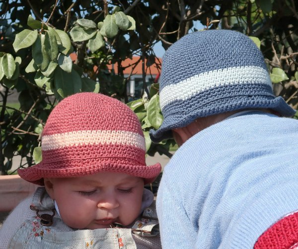 A baby and a toddler in a garden wearing crochet sun hats.