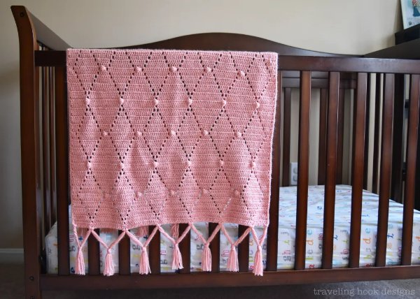 Pink filet crochet baby blanket hanging over a crib.
