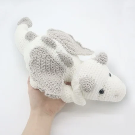 A white and pale grey crochet dragon sitting on someones hand.