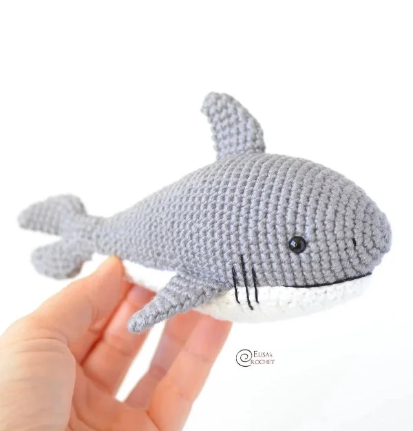 A grey and white crochet shark being held in someones hand.