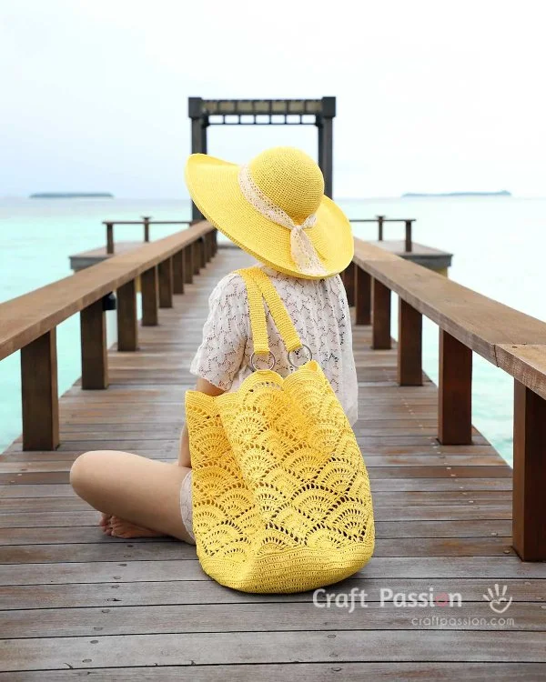 A woman sitting on a jetty over the water with a bright yellow hat and crochet beach bag.