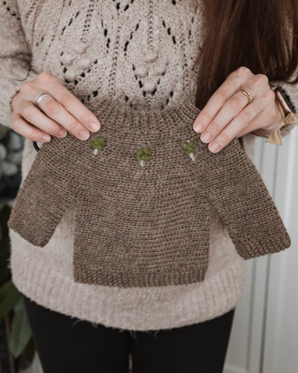 A woman holding a tiny crochet baby sweater with little embroidered tree detail.