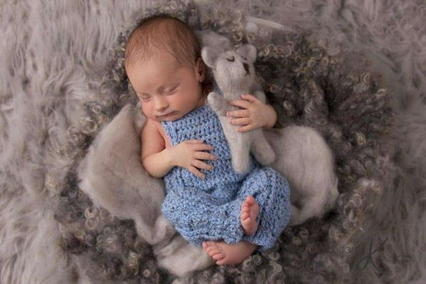 A newborn baby in blue crochet overalls with a toy and fur.