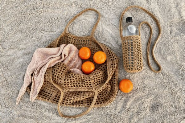A natural colourec crochet beach bag on the sand with oranges and a water bottle.