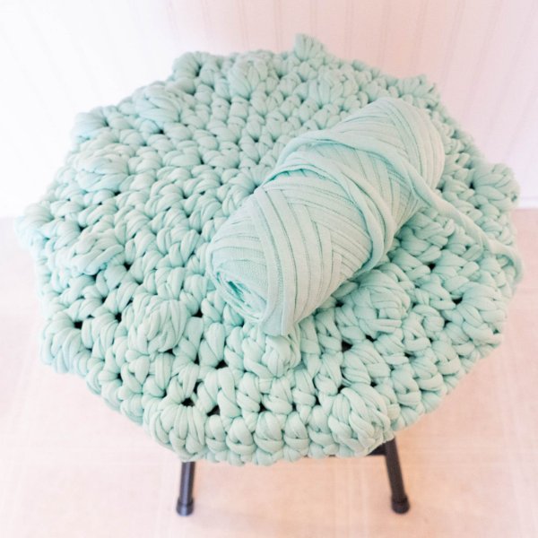 A closeup of a stool with a pale blue crochet stool cover and a ball of yarn.