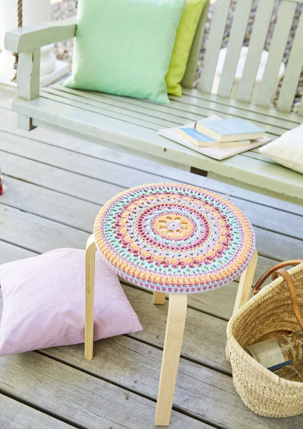 A crochet stool cover on a foot stool on a verandah with cushions and a bench.