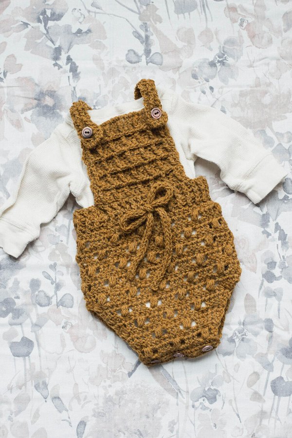 A mustard coloured, bobble stitch crochet baby romper with buttons and a bow.