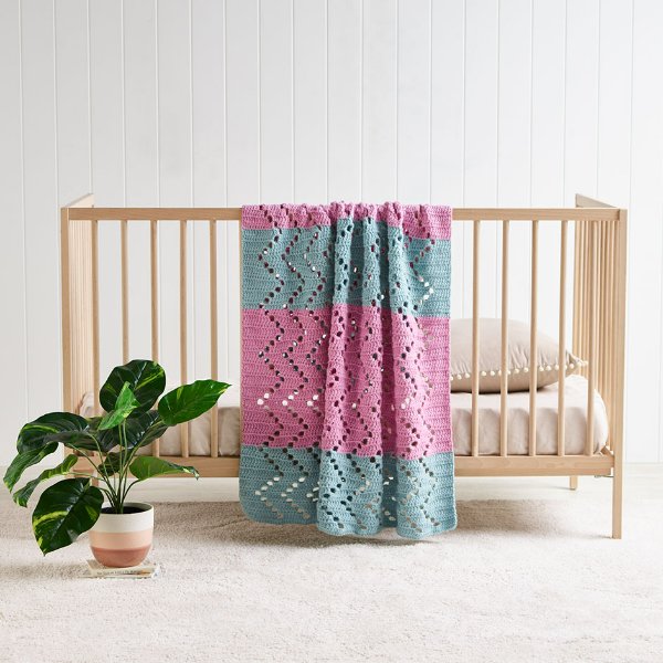A pink and blue filet crochet baby blanket draped over a timber crib.