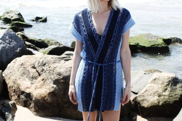 A woman on a rocky beach weraing a blue crocheted beach robe.