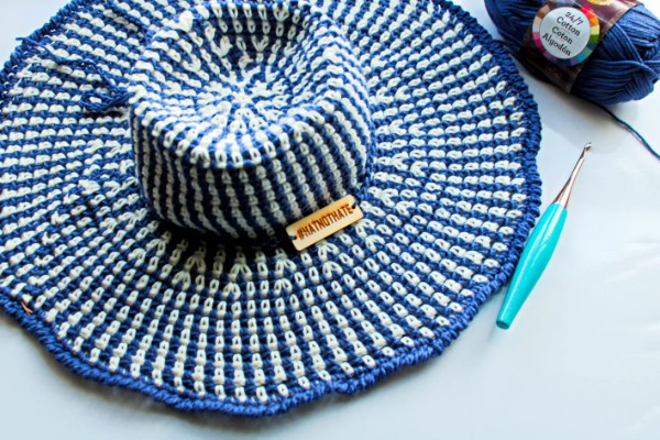 A blue and white wide-brimmed crochet sun hat with a ball of yarn and a crochet hook.