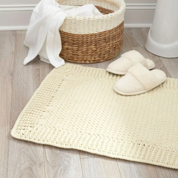 A cream coloured crochet bath mat in a bathroom with slippers and a linen basket.