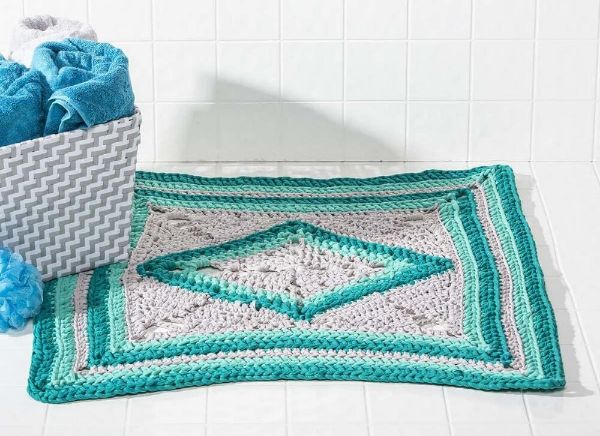 A green and greige crochet bath mat on white timber boards.