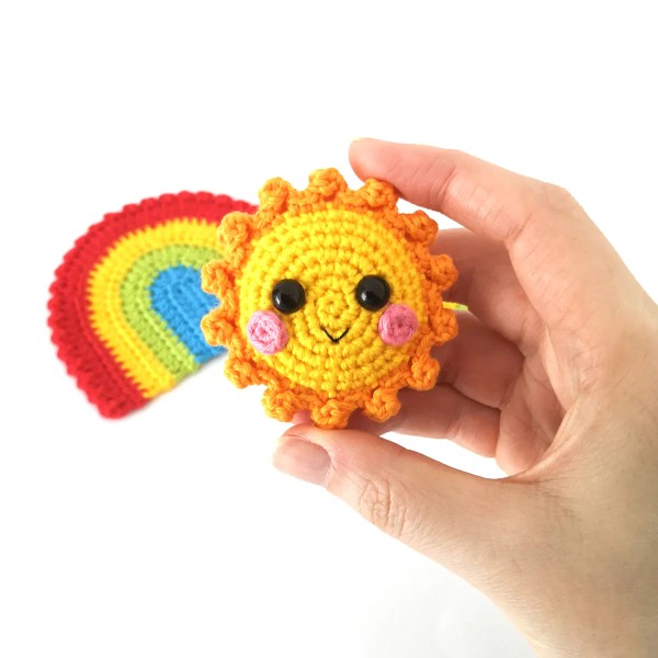 A crochet sun keychain with a crochet rainbow in the background,