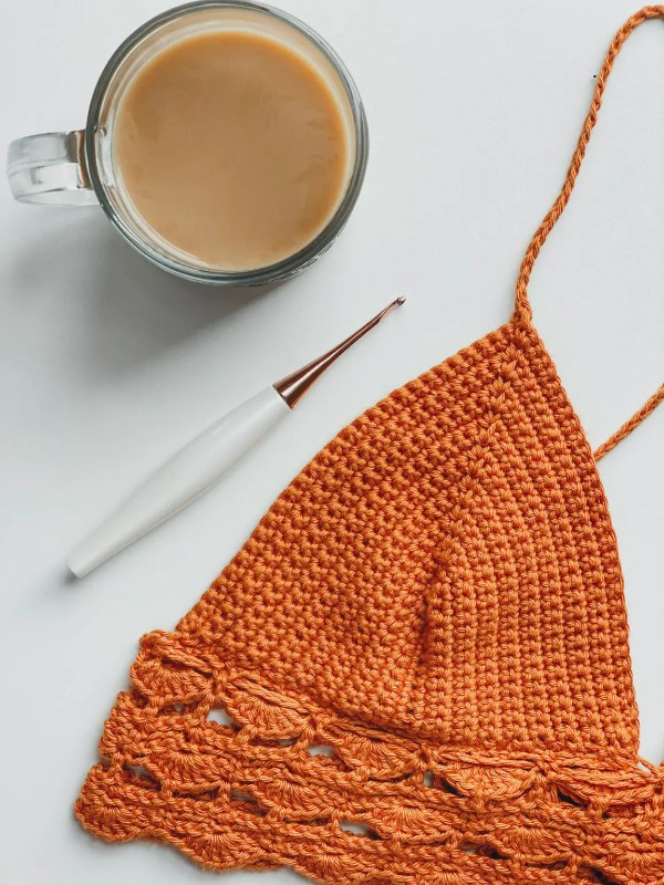 A closeup of an orange crochet bralette, a crochet hook and a cup of coffee.