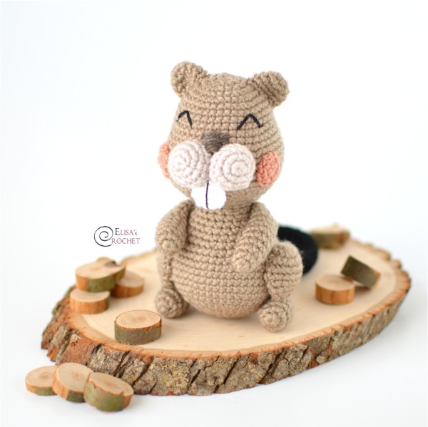 A crochet beaver with pink cheeks on a wooden log.