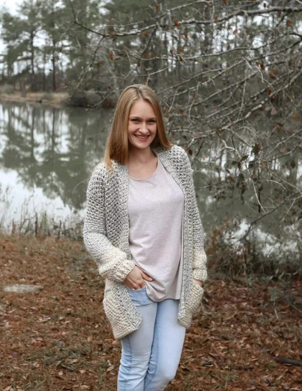 A woman by a lake wearing a pale grey and blue crochet cardigan with pockets.