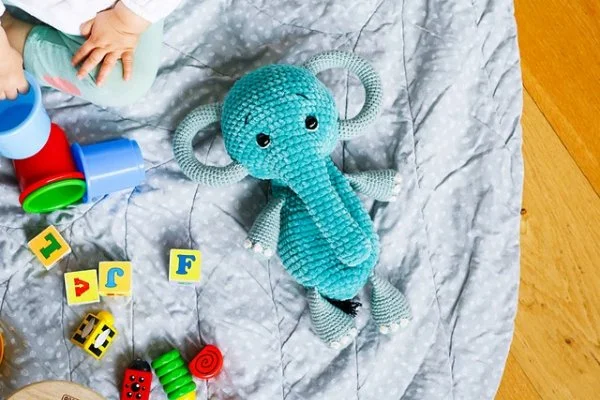 A blue crochet elephant on a baby playmat surrounded by toys.