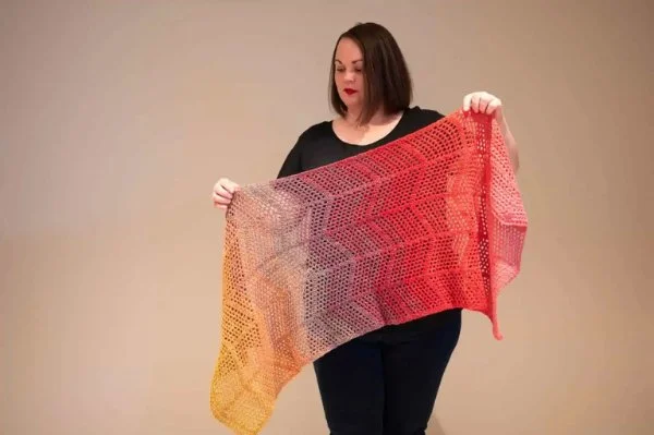 A woman holding a large, lacy crochet scarf worked in gradient yarn.