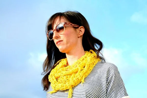 A woman wearing a bright yelllow lacy crochet scarf with a bright blue sky background.