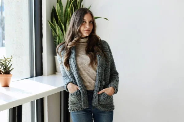A woman weraing a grey coloured crochet cardigan with a rolled collar and pockets.